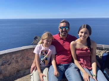 A man and two girls sit on a stone ledge with a stunning blue ocean behind them. All are smiling, enjoying the sunny, scenic day.
