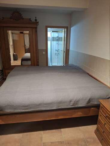 This image shows a bedroom with a large wooden bed, grey bedding, tiled floor, antique wardrobe with mirror, and an ensuite bathroom featuring a glass shower.