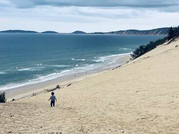 Ein Kind läuft eine steile Sanddüne runter in Richtung eines riesigen, ruhigen Ozeans unter einem bewölkten Himmel, mit fernen Hügeln und sanften Wellen, die das friedliche Bild abrunden.