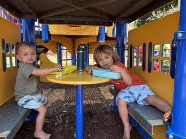 Zwei kleine Kinder sitzen an einem gelben Tisch unter einem Spielgerät, essen Snacks aus ihren Lunchboxen und genießen die Zeit zusammen an einem sonnigen Tag.