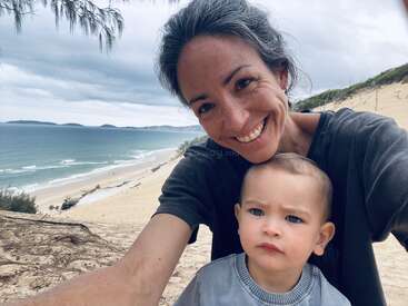 Eine Frau und ein kleines Kind machen ein Selfie auf einem sandigen Hügel mit Blick aufs Meer, mit Wellen, bewölktem Himmel und fernen Inseln im Hintergrund.