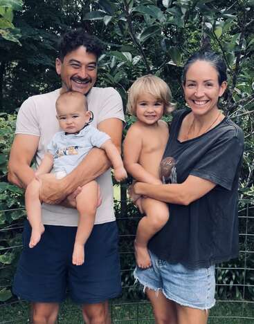 Eine fröhliche vierköpfige Familie steht draußen, die Eltern halten jeweils ein kleines Kind im Arm, alle lächeln glücklich. Üppiges Grün und Bäume bilden einen lebendigen, natürlichen Hintergrund.