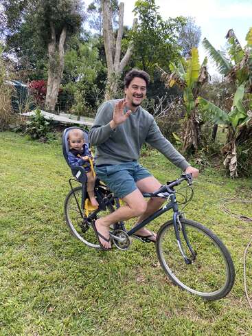 Ein lächelnder Typ fährt mit dem Fahrrad über Gras und winkt in die Kamera. Ein Baby sitzt hinter ihm in einem Kindersitz, umgeben von grünen Bäumen und Pflanzen.