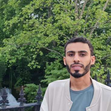 A young man with a trimmed beard stands in front of a lush green forest and metal fence. A waterfall is visible in the background, creating tranquility.