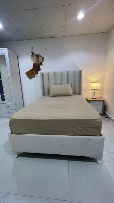 A neatly made single bed with beige sheets, a matching pillow, a lamp on a nightstand, a wardrobe, and damaged wall paint behind the bed.