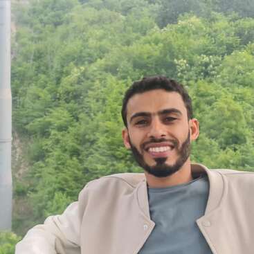 A smiling man with a trimmed beard sits relaxed, wearing a grey shirt and beige jacket, with a lush green forest and hills in the background.