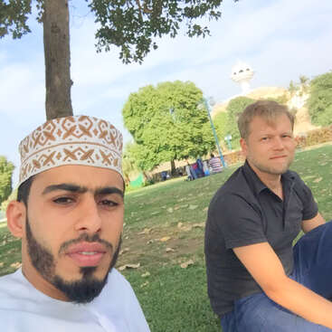 A collage of selfies featuring one man in traditional Omani attire with various friends, outdoors in parks, by the sea, at landmarks, and near a car.
