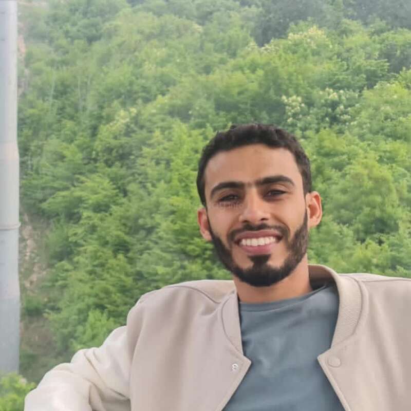 A smiling man with a trimmed beard sits relaxed, wearing a grey shirt and beige jacket, with a lush green forest and hills in the background.