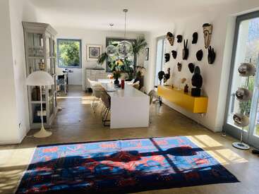 Bright dining area with large windows, modern white table, eclectic chairs, colorful rug, wall-mounted African masks, glass cabinets, and lush plants, blending contemporary and ethnic styles.