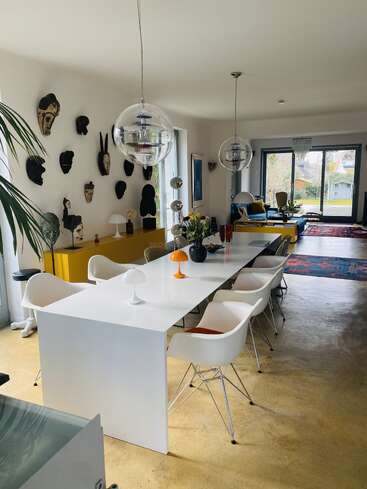 A modern dining and living area with a long white table, designer chairs, eclectic wall masks, pendant lights, yellow sideboard, and large windows overlooking greenery.