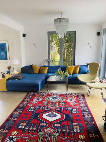 A modern living room with a blue sectional sofa, yellow pillows, glass coffee table, red patterned rug, chandelier, artwork, and large windows letting in natural light.