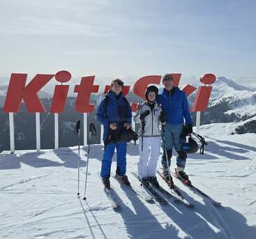 Three people in ski gear stand happily on a snowy mountain, posing in front of a large red "KitzSki" sign with stunning alpine scenery behind.