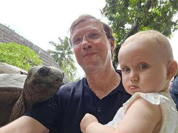 A man holding a baby poses for a photo with a large tortoise outdoors. Green trees, bushes, and a bright sky are visible in the background.