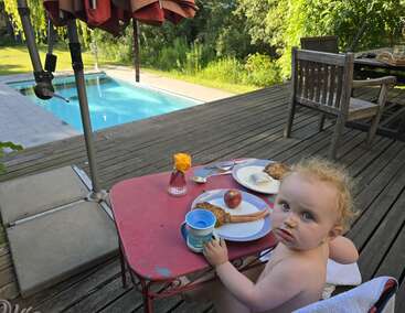 A toddler sits at a small red table outdoors, eating breakfast next to a swimming pool. Sunlight, greenery, wooden deck, and a flower vase complete the scene.