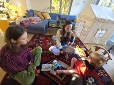 Three children sit on a colorful carpet in a cozy living room, playing with toys. There is a couch, play tent, and Christmas tree in the background.