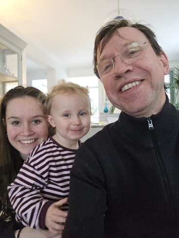 A smiling man, a woman, and a young child pose happily for a selfie indoors. The atmosphere is warm, bright, and filled with joy and love.