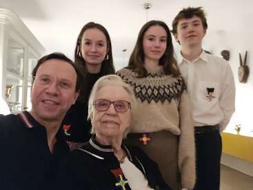 A family of five, spanning generations, poses together indoors. All wear medals pinned to their clothing and smile warmly at the camera, creating a celebratory atmosphere.