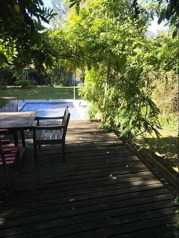 A wooden deck with chairs and table is shaded by lush green trees. Beyond the deck, a pool is covered and grassy yard stretches out.