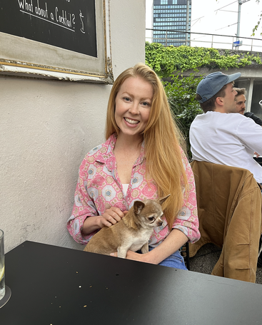 Une femme souriante aux longs cheveux roux est assise à une table noire, tenant un petit chihuahua marron sur ses genoux, en plein air, dans un café confortable.