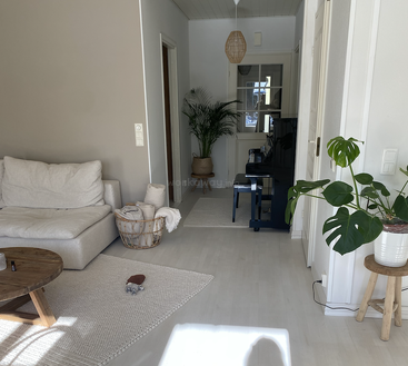 Un salon confortable et minimaliste est composé d'un canapé beige, d'une table basse en bois, de tapis clairs, de plantes vertes luxuriantes, d'un panier avec des couvertures et d'un piano noir brillant.