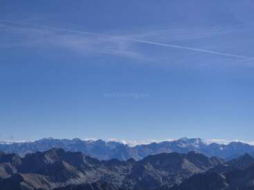 Jagged mountain peaks stretch across the landscape under a vast, clear blue sky. Distant snow-capped summits glisten, while a single jet trail arcs above. Serene beauty.