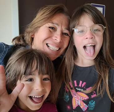 A joyful older woman and two young girls pose for a selfie, smiling and making funny faces; the girl on the right sticks out her tongue playfully.