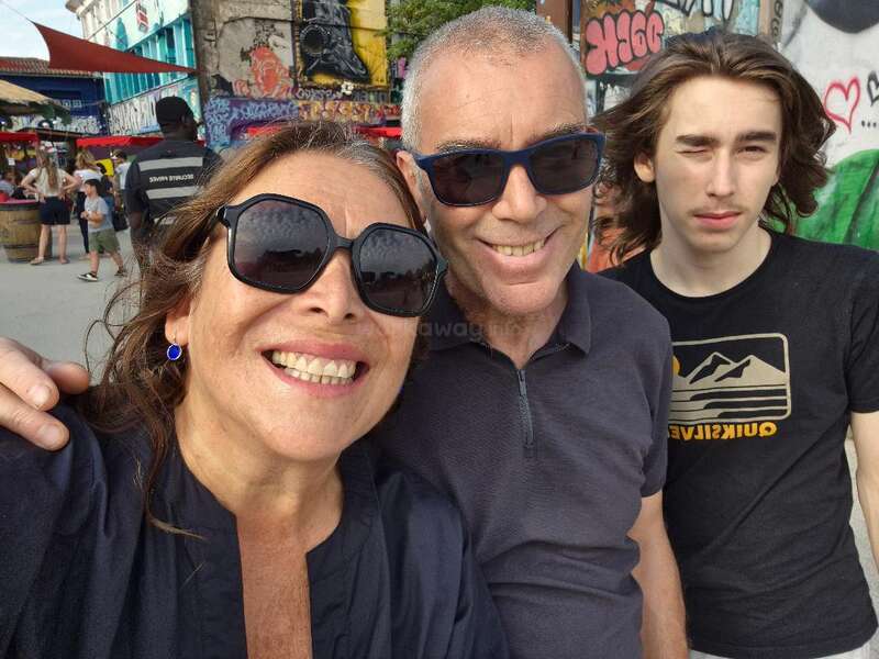 Three people smile for a selfie outdoors, wearing sunglasses. Behind them, colorful graffiti decorates the walls. Other people wander around, creating a lively urban atmosphere.