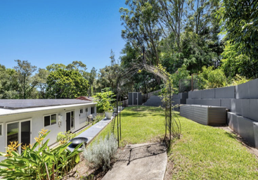 Cette image montre une arrière-cour ensoleillée avec une pelouse luxuriante, des parterres de jardin, un chemin pavé, de la verdure, des murs de soutènement, un abri et une maison blanche.