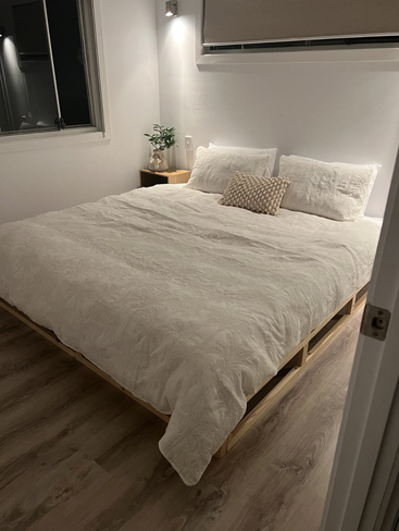 Une chambre à coucher minimaliste et confortable avec un grand lit recouvert de draps blancs, deux oreillers blancs, un coussin décoratif, une table de chevet, une plante et un éclairage doux.
