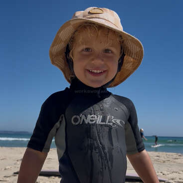 Un jeune enfant souriant, vêtu d'une combinaison de plongée et d'un chapeau de soleil, se tient sur une plage ensoleillée au ciel bleu. L'océan et les surfeurs sont visibles à l'arrière-plan.