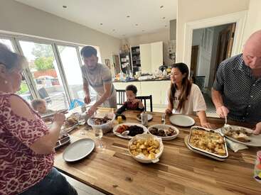 A family gathers around a wooden table filled with delicious food, sharing a meal together in a bright, cozy kitchen with large windows and sunlight.