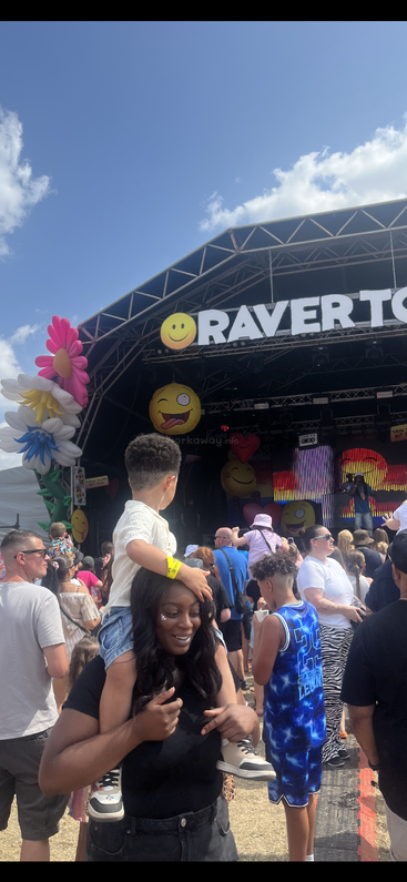 A lively outdoor music festival with colorful decorations, big emoji faces, happy attendees, a woman carrying a child on her shoulders, and a sunny blue sky.