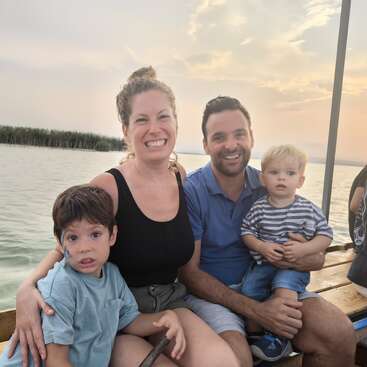 Una sonriente familia de cuatro miembros se sienta junta en un banco de madera junto al agua al atardecer, disfrutando de un tranquilo y alegre paseo en barca al aire libre.