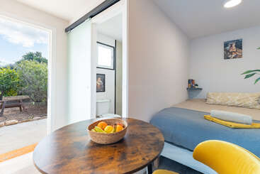 This image depicts a cozy bedroom with a bed, small dining table with a fruit bowl, a bathroom, open door to a garden, and sunlight.