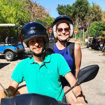 The image depicts a man and woman riding a scooter, both wearing helmets, with the man in a green shirt and the woman in a blue tank top, set against a scenic background.