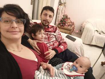 A family of four sits together on a cozy couch. Smiling parents, two young children, and a decorated Christmas tree create a warm, festive atmosphere.