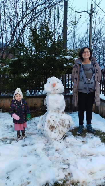 A smiling woman and a young girl stand outside in the snow beside a snowman with a carrot nose and stick arms, surrounded by winter scenery.