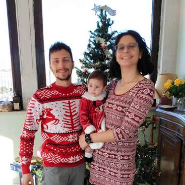 A smiling family stands by a decorated Christmas tree. Both adults wear festive sweaters, while the baby is dressed in a cute red holiday outfit. Cozy atmosphere.