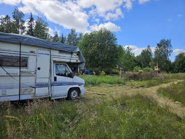 Un camping-car blanc est garé sur un terrain herbeux entouré d'arbres. Un chemin de terre serpente à proximité sous un ciel bleu parsemé de nuages. Une scène paisible.