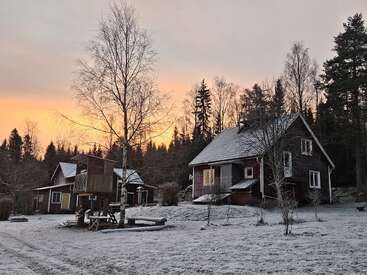 Une maison rouge confortable se trouve dans une cour enneigée, entourée d'arbres au lever du soleil. Des structures de jeux et des dépendances ajoutent du charme à la forêt hivernale en arrière-plan.