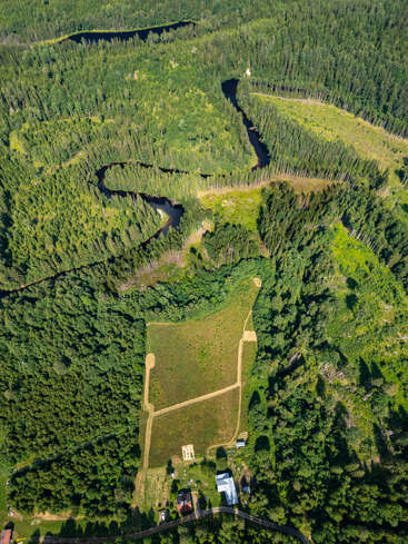 Une rivière sinueuse serpente à travers une forêt dense et verdoyante. En contrebas, une clairière rectangulaire abrite quelques bâtiments. La lumière du soleil met en valeur les arbres luxuriants et les espaces verts.