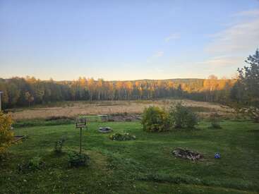 A peaceful rural landscape at sunrise, featuring a green garden, bird feeder, fire pit, open field, distant forest, and hills beneath a clear blue sky.