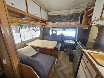 This cozy camper interior features a dining table, cushioned seating, kitchenette with sink, stove, fridge, overhead storage, bunting, and an upper sleeping area above the cab.
