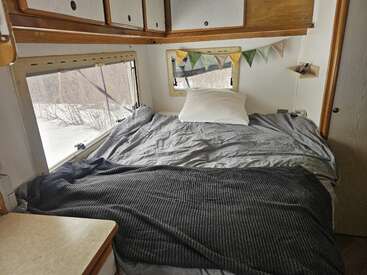 A cozy bed with gray bedding inside a small camper. Large windows provide a snowy view. Cabinets, bunting, and a soft pillow add comfort and charm.