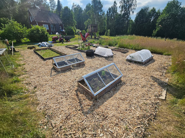 A sunny garden with raised beds, some covered with glass and mesh, surrounded by wood chips, bordered by grass, trees, a red house, and various equipment.