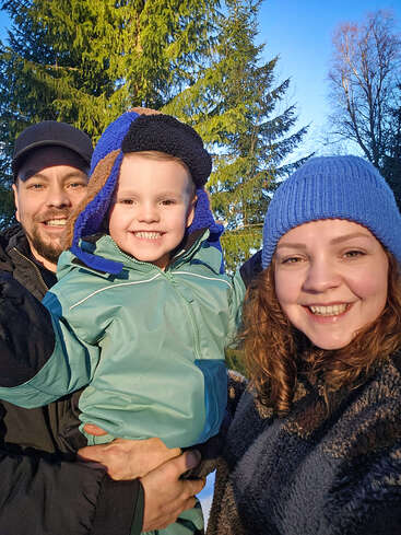 A smiling family of three poses outdoors in winter clothing. The child, in a blue jacket and hat, is held between two happy adults under bright sunshine.