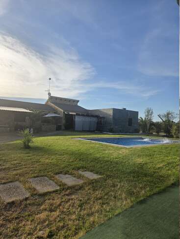 Uma casa moderna de pedra fica sob um céu azul, cercada por grama verde. Tem uma piscina retangular e pedras que levam até o prédio. Um clima tranquilo.