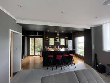A modern kitchen with dark walls, black countertops, and three bar stools with red seats. Large windows provide natural light and scenic outdoor views. Cozy atmosphere.