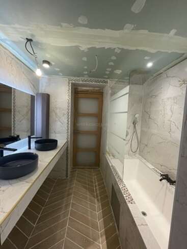 This image shows a modern bathroom under renovation, featuring two dark sinks, a large mirror, bathtub with a shower, tiled floor, and unfinished ceiling.
