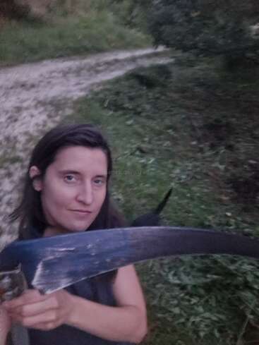 Une femme se tient à l'extérieur sur un chemin herbeux, tenant une grande faux. L'arrière-plan est vert avec des arbres et l'éclairage suggère le début de la soirée ou le crépuscule.